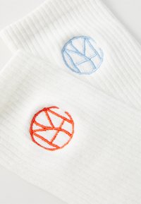 Dos calcetines blancos acanalados con diseños circulares bordados contrastantes; uno en azul, el otro en naranja. Textura suave y patrón simple.