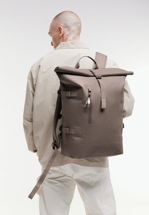 Muž s tetováním, oblečený v béžové bundě a kalhotách, nese velký taupe batoh s rolovacím uzávěrem, bočními popruhy a přední kapsou na zip.