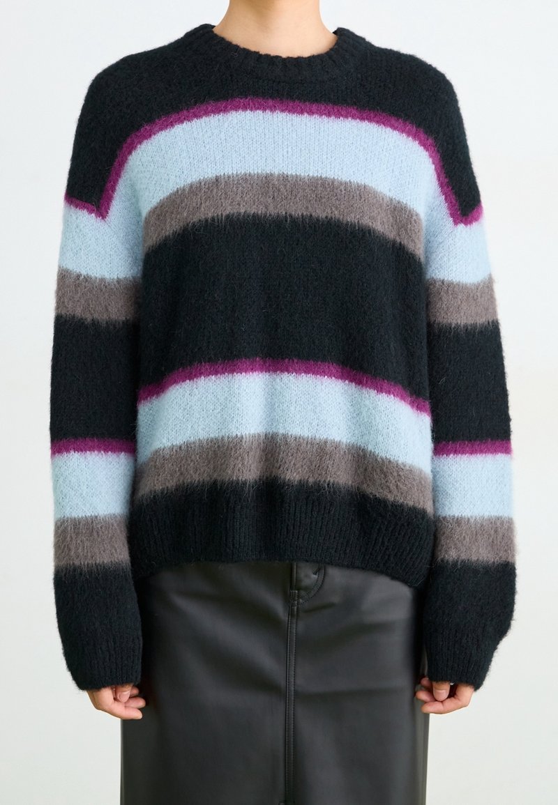 Personne portant un pull noir duveteux avec des rayures horizontales bleu clair, grises et violettes, assorti à une jupe en cuir noire.