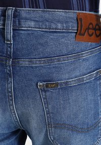 Blue denim jeans with a brown leather label featuring "Lee" on the waistband. The fabric has a classic straight cut and textured finish.