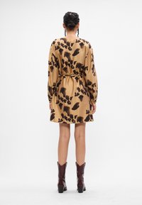 Καφέ φόρεμα με μαύρα αφηρημένα σχέδια, μακριά μανίκια, λεπτή μέση και φαρδιά μέση. Φορεμένο με καφέ μποτάκια μέχρι τον αστράγαλο. Απλή άσπρη φόντο.