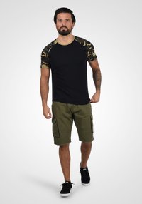 T-shirt noire avec des manches camouflage, associée à un short cargo vert olive. Tissu texturé, encolure ronde et baskets noires.