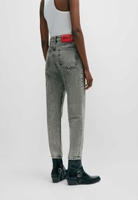 Harmaat korkeavyötäröiset farkut, joissa on haalistunut pinta, takataskut ja punainen "HUGO" -merkki; yhdistetty mustiin nilkkabootseihin, joissa on solki.