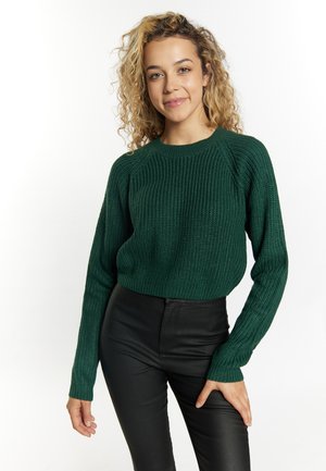 Jonge vrouw met krullend blond haar, dragend een donkergroene gebreide trui en zwarte leren broek, staand tegen een witte achtergrond.