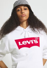 Felpa bianca con logo rosso Levi's di grandi dimensioni sul petto, realizzata in tessuto morbido con cappuccio regolabile. Abbinata a un cappello a secchiello a scacchi bianco e nero.
