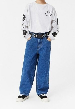 Niño con camisa blanca de manga larga con texto en círculo negro, vaqueros anchos azules, cinturón negro y zapatillas blancas y negras, con las manos en los bolsillos.