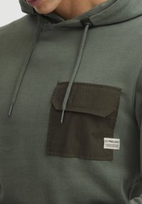 Green cotton hoodie featuring a textured dark green chest pocket, adjustable drawstrings, and small label detailing the brand.