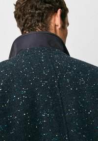 Mörk tweedkappa med en herringbone-mönster, med en hög krage som har en kontrasterande marinblå foder. Texturen verkar mjuk och något grov.