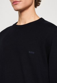 Jeune personne portant un pull noir à col rond avec "BOSS" brodé sur la poitrine, sur un fond blanc uni.