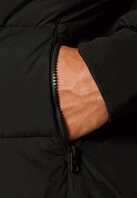 Sort dunjakke med lynlåslomme, der viser et nærbillede af en hånd, der griber lynlåsen. Glat tekstur, minimalistiske designelementer.