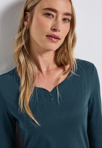 Haut à manches longues teal avec un col rond en V, deux boutons à l'avant, et une texture de tissu lisse et douce.