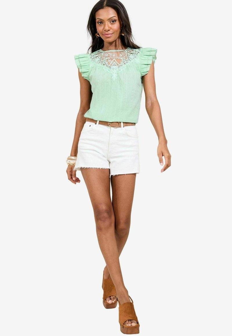 Blusa verde menta con maniche a volant e dettagli in pizzo, abbinata a shorts in denim bianchi e sandali con tacco color nude. Design semplice e strutturato.