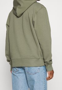 Felpa verde oliva realizzata in misto cotone, con cappuccio regolabile, polsini e orlo a costine, abbinata a jeans azzurri chiari.