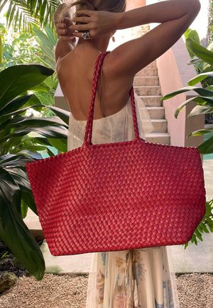 Frau, die mit erhobenen Armen ihre Haare richtet, trägt eine große rote gewebte Tragetasche und steht in der Nähe tropischer Pflanzen und steinerner Treppen.