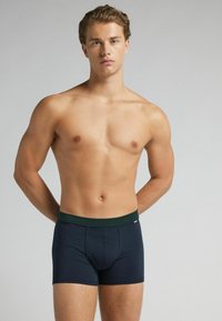 Ung man med ljus hy och lockigt hår iförd marinblå boxershorts som står mot en ljus bakgrund.