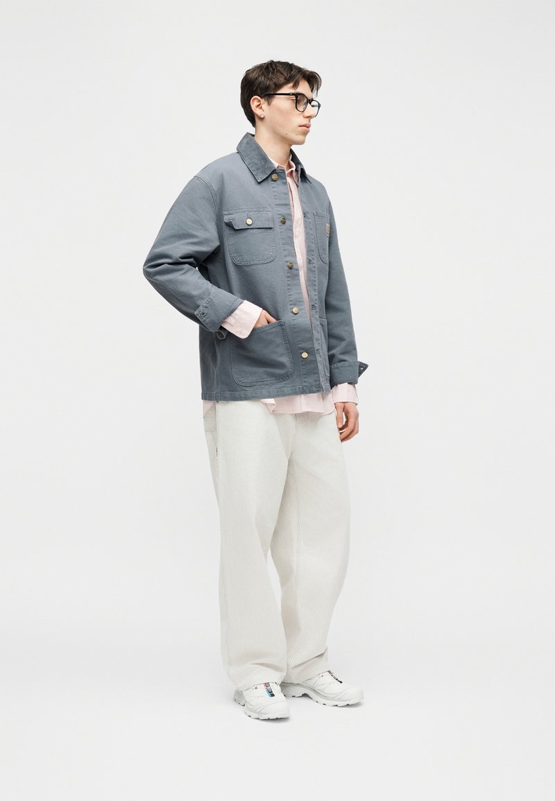 Giovane uomo con capelli corti e occhiali che indossa una giacca grigia sbottonata, camicia rosa chiaro, pantaloni bianchi larghi e sneakers bianche, in piedi con una mano in tasca.