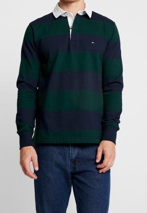 Mężczyzna w długim rękawie koszulki rugby w paski w kolorze ciemnej zieleni i granatu, z białym kołnierzem i małym logo na piersi, w połączeniu z niebieskimi dżinsami.