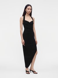 Γυναίκα που φοράει μαύρο φόρεμα με halter neck και ασύμμετρο τελείωμα και μαύρα ανοιχτά πέδιλα με τακούνι στέκεται μπροστά σε λευκό φόντο.