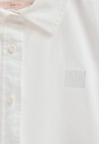White slim-fit shirt featuring a classic collar, button closure, and a stitched logo patch. Smooth cotton fabric with subtle texture.