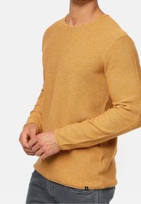 Suéter de manga larga con cuello redondo en color mostaza, hecho de un tejido de punto texturizado. Tiene un corte relajado y detalles de costura sutiles a lo largo de las costuras.