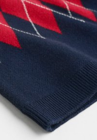 Gros plan sur une manche de pull bleu marine avec un motif argyle rouge et blanc et une bordure côtelée sur une surface blanche.