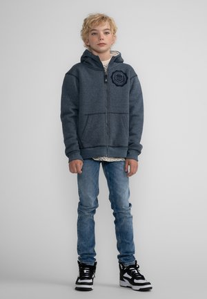 Felpa con cappuccio e zip in tessuto grigio scuro, con tasche frontali ed emblemma ricamato. Indossata sopra una camicia a fantasia e abbinata a jeans blu e sneakers.