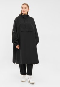 Black long-sleeve waterproof coat with a high collar, a front pocket, and elastic cuffs, worn over black pants and sneakers.