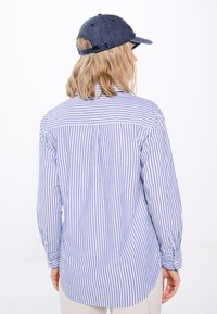 Camicia a righe blu e bianche con bottoni e maniche lunghe, orlo curvo e piega sul retro, abbinata a un cappellino scuro.