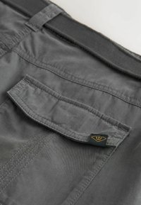 Pantalon en tissu gris avec une poche arrière à rabat et une petite étiquette noire ornée d'un logo doré en forme de trident près de la taille.