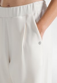 Weiße, plissierte Hose aus einem weichen, glatten Material mit elastischem Bund, seitlichen Taschen und einem kleinen grauen Branding-Detail auf der Tasche.