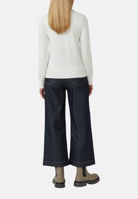 Un pull en tricot blanc avec un ourlet côtelé, associé à un jean large bleu foncé et des bottines beiges. La tenue est vue de l'arrière.