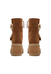 Botines de ante marrón con tacón grueso y acanalado, correas ajustables y detalles de hebillas doradas. Cierre trasero con cremallera para facilitar su uso.