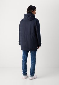 Navy blue jacket with a hood, featuring a straight cut, side pockets, and adjustable cuffs. Paired with blue jeans and white sneakers.