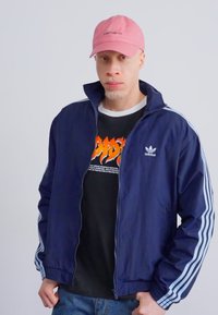 Jūrinės sportinės striukės su baltomis juostelėmis, užtrauktuku ir "Adidas" logotipu; juoda grafika marškiniai po ja; rožinė kepurė. Lygus fonas.