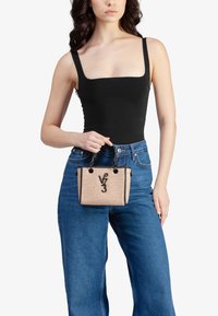 Donna che indossa un top senza maniche nero e jeans blu, mentre tiene una piccola borsa beige intrecciata con manici neri e un logo "V 3".