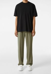 Muž v černém oversized tričku s krátkým rukávem, volných olivově zelených kalhotách a bílých teniskách, stojící na pozadí čistě bílé stěny.