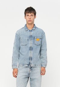 Light blue denim jacket with long sleeves, front zipper, two chest pockets, and a small orange label on the left pocket. Smooth texture.