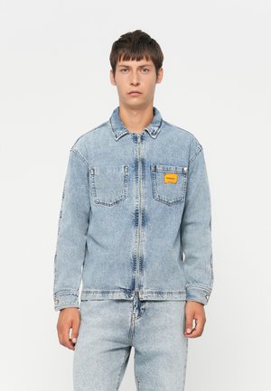 Light blue denim jacket with long sleeves, front zipper, two chest pockets, and a small orange label on the left pocket. Smooth texture.