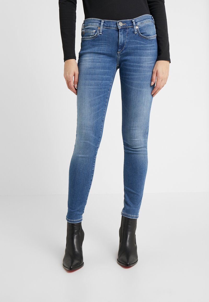 Kapeje siniset skinny farkut keskimääräisellä pesulla, joissa on nappisulku, etutaskut ja minimaalinen kuluminen, yhdistettynä mustiin nilkkabootseihin.