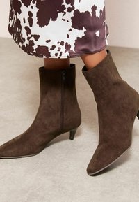 Rudos spalvos zomšiniai kulkšnies batai su smailiu noseliu ir žemu kulnu, turintys šoninį užtrauktuką patogiam dėvėjimui. Lygus paviršius ir švarus siluetas.