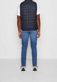 Mörkblå vadderad puffärmelväst med hög krage, designad för lager-på-lager, tillsammans med blå jeans och grå skor, sedd bakifrån.