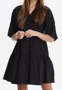 Femme portant une robe noire ample à manches bouffantes courtes et jupe à volants, se tenant devant un fond blanc uni.