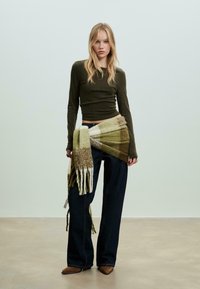 Mørkegrøn tætsiddende langærmet bluse, brede mørke jeans og et struktureret tørklæde med grønne og brune striber om livet.