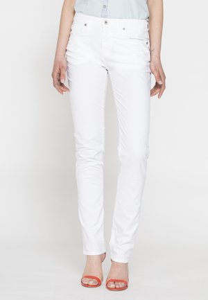 Persona che indossa pantaloni bianchi slim e sandali arancioni con tacco alto e punta aperta, in piedi contro uno sfondo chiaro e uniforme.