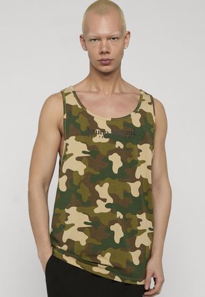 Ung person med kort blondt hår iført en grønn og beige kamuflasjetanktopp og svarte bukser, stående mot en ensfarget bakgrunn.