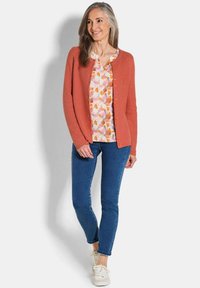 Cardigan corail avec un tricot texturé, encolure ronde et boutons oranges, superposé sur une blouse florale, associé à un jean bleu et des baskets.