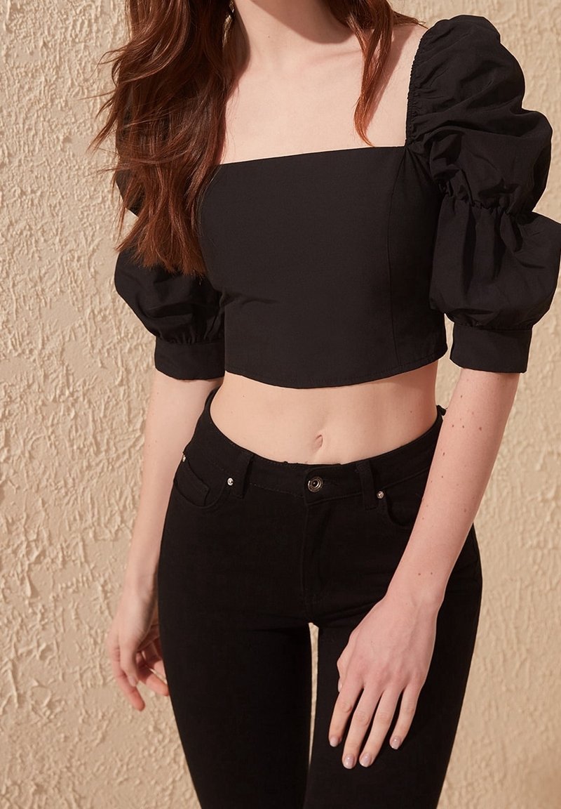 Top corto nero con scollo quadrato e maniche a palloncino, abbinato a jeans neri a vita alta. Tessuto morbido, design aderente, texture lucida sottile.