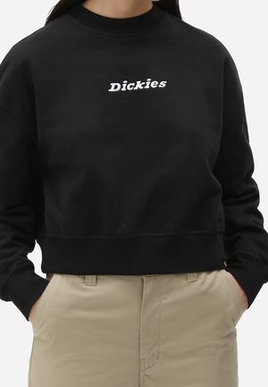 Persoon draagt een zwarte cropped sweatshirt met de tekst "Dickies" in het wit, handen in de zakken van beige broek.
