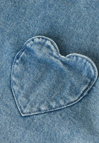 Džinsa audums ar gaiši zilu sirds formas ielāpu, izšūtu ar redzamu diegu, uz teksturēta zilā fona.