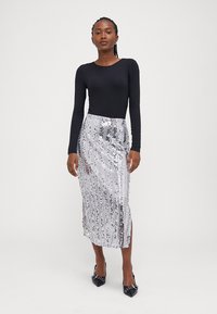 Top negru cu mânecă lungă purtat cu o fustă midi argintie cu paiete, prezentând dungi verticale și o fente laterală; modelul poartă tocuri negre decorate.